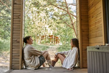 Dışarıdaki ahşap yazlığın verandasında şarabın yanında oturan pozitif romantik çiftin yan görüntüsü.