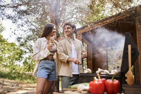 stock image Smiling woman holding wine and standing near boyfriend, bbq with vacation house at background