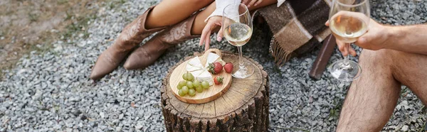 stock image Cropped view of couple holding wine near fresh fruits and cheese outdoors during picnic, banner