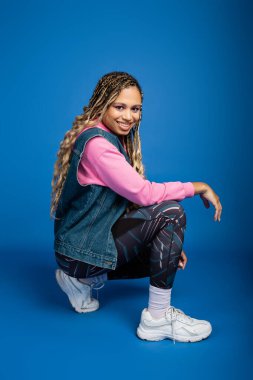 Mavi arka planda rastalı poz veren mutlu Afro-Amerikan kadın, spor kıyafet, gülümseme