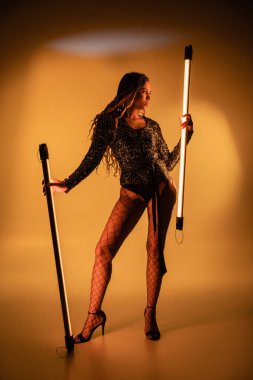 Seksi, Afro-Amerikalı kadın sarı arka planda floresan lambalar tutuyor, yüksek topuklu.