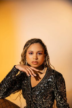 Parti kıyafetli güzel bir kadın sarı arka planda kameraya bakıyor, koyu tenli bir model.