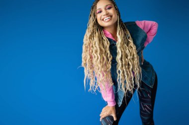 Mavi arka planda poz veren pembe kapüşonlu ve kot ceketli mutlu Afro-Amerikan kadını