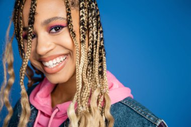 Çekici Afro-Amerikalı kadın pembe kapüşonlu ve kot ceketli kameraya bakıyor, mavi arka plan