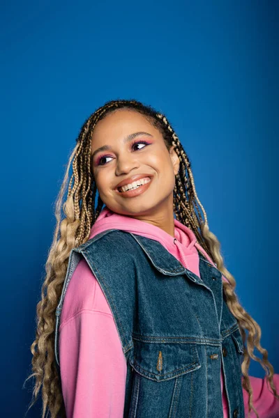 happy african american woman with dreadlocks and pink hoodie smiling and looking away on blue
