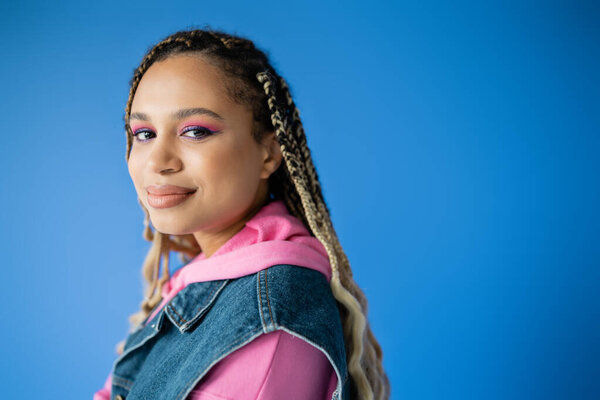 happy african american woman with dreadlocks looking at camera on blue background, bold makeup