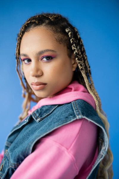attractive african american woman with dreadlocks looking at camera on blue background, bold makeup