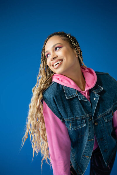 joyful african american woman in pink hoodie and denim vest smiling on blue background, positive