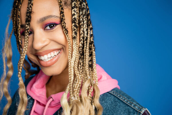 attractive african american woman in pink hoodie and denim vest looking at camera, blue background