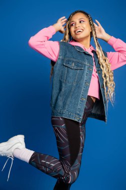 Koyu tenli, spor kıyafetli, mavi arka planda gülümseyen, cesur makyajlı, mutlu bir model.