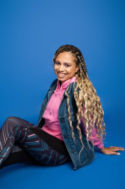 Uzun saçlı mutlu Afro-Amerikan kadın kameraya bakıyor, mavi arka planda oturuyor, sportif.