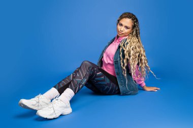 full length of long haired african american woman in sporty outfit sitting on blue background clipart