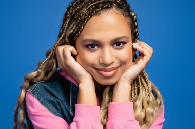 Çekici ve mutlu Afrikalı Amerikalı kadın mavi arka plandaki kameraya bakıyor, moda ve güzellik.