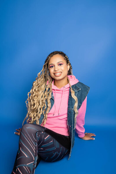 happy african american woman in sporty outfit sitting on blue background, looking at camera