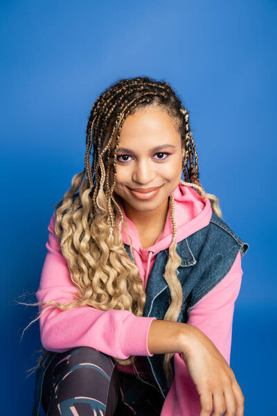 dark skinned woman in pink hoodie, denim vest and leggings looking at camera on blue background