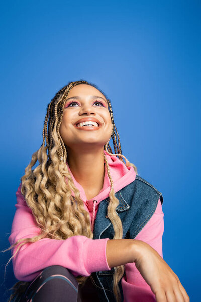 happy dark skinned woman in pink hoodie and denim vest looking up while sitting on blue background