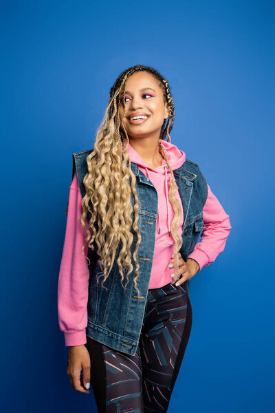 happy african american woman with dreadlocks standing with hand on hip on blue background, studio