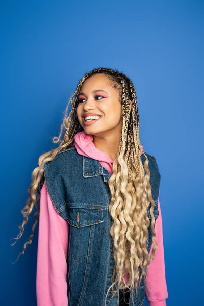 joyful african american woman with dreadlocks smiling on blue background, denim vest and hoodie