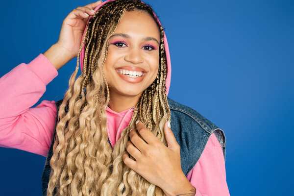 positive and pretty african american woman with hood on head smiling on blue background, denim vest