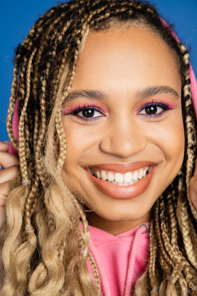 close up of cheerful black woman with hood on head looking at camera and smiling on blue background