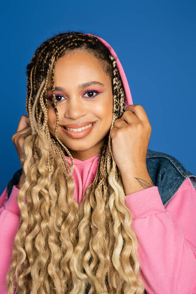positive dark skinned woman with hood on head looking at camera and smiling on blue background