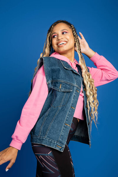positive dark skinned woman with dreadlocks looking away, blue background, pink hoodie, denim vest