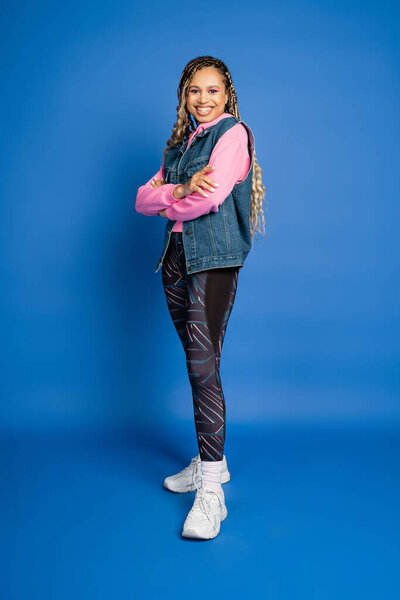 full length, dark skinned woman in sporty outfit posing with folded arms on blue background, happy