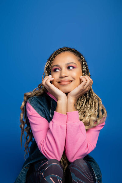 charming and dreamy african american woman with bold makeup looking away on blue background