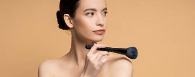 Asian woman with perfect skin holding cosmetic brush near naked shoulder isolated on beige, banner clipart