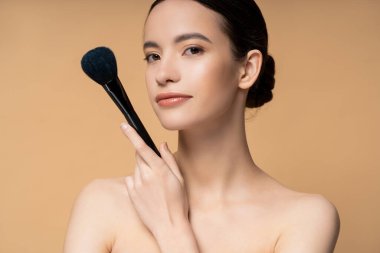 Confident young asian woman with naked shoulders holding makeup brush and standing isolated on beige clipart