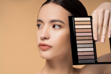 Portrait of young asian woman with natural makeup holding eyeshadow palette isolated on beige clipart