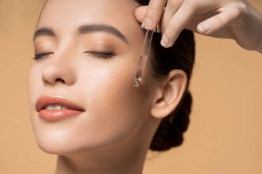 Memnun Asyalı kadının bej üzerinde tek başına dikilirken kozmetik serumu uygulamasını yakından izle