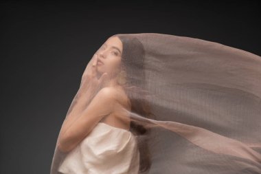 Esmer Asyalı model çeneye dokunuyor ve bej kumaşın altında poz veriyor.