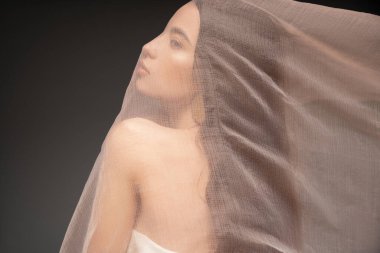 Side view of asian model with naked shoulder looking away while posing under cloth isolated on black clipart