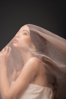Asyalı, uzun saçlı, çıplak omuzlu bir kadın bej kumaşın altına bakıyor.
