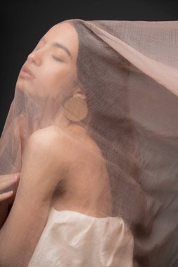 Üzerinde güzel bir Asyalı model ve küpe bej kumaşın altında poz veriyor.