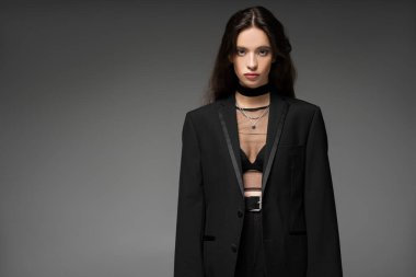 Ceketli ve gümüş zincirli şık Asyalı model gri üzerinde tek başına dururken kameraya bakıyor.