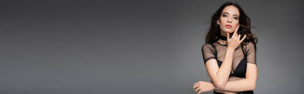 Attractive asian woman in mesh top and necklaces looking at camera isolated on grey, banner