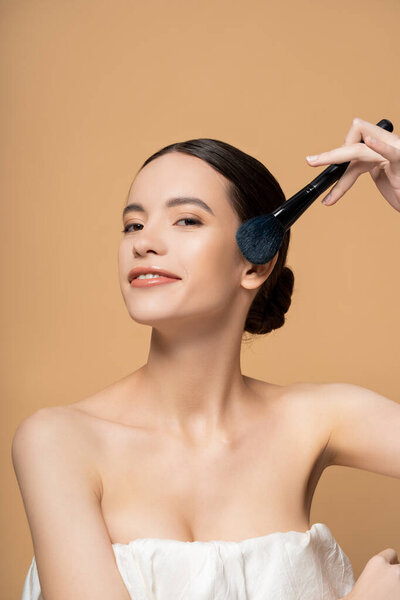 Улыбающаяся молодая азиатская женщина с обнаженными плечами, держащая румяную щетку возле щеки, изолированной на бежевом