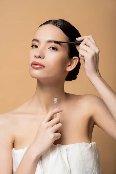 stock image Confident young asian woman in top applying eyebrow gel and looking at camera isolated on beige
