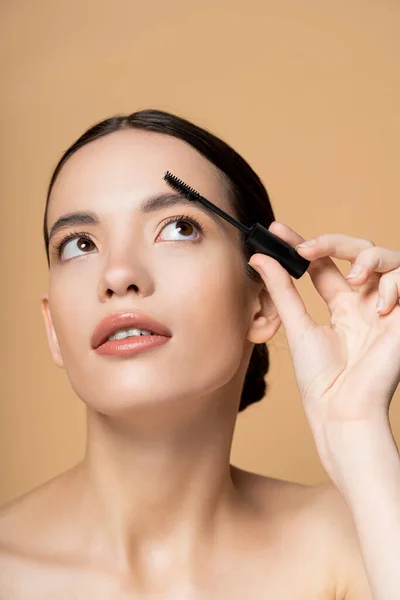 stock image Pretty young asian woman with naked shoulders looking up and apply eyebrow gel isolated on beige