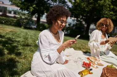 Afrika kökenli Amerikalı bir kadın yaz pikniğinde kız arkadaşıyla akıllı telefon kullanıyor.