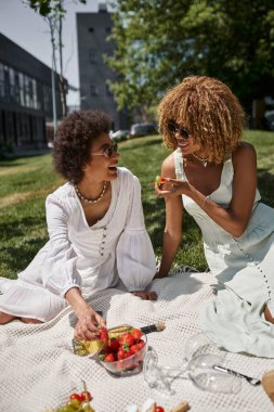 joyful african american women eating fruits and chatting on summer picnic in park clipart