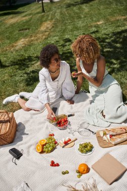 Gülen Afrikalı Amerikalı kız arkadaşlar meyve yiyor ve parkta piknikte battaniyenin üzerinde konuşuyorlar.