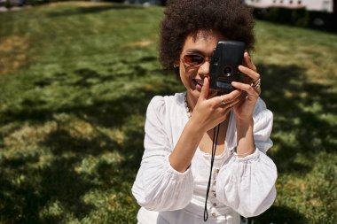Güneş gözlüklü mutlu bir Afro-Amerikan kadını yaz parkında fotoğraf çekiyor.
