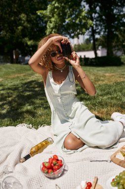 Genç Afro-Amerikalı kadın, yaz parkında piknik yaparken klasik kamerayla fotoğraf çekiyor.