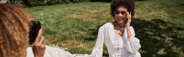 Eski model fotoğraf makinesi olan Afro-Amerikan kadın yaz parkında mutlu kız arkadaşının fotoğrafını çekiyor.
