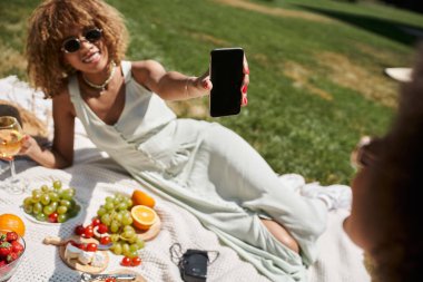 Gülümseyen Afrikalı Amerikalı kadın yaz parkında piknik yaparken elinde boş ekranla akıllı telefon tutuyordu.