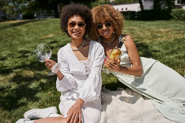 Piknik keyfi, kaygısız Afrikalı Amerikalı kız arkadaşlar, bir şişe şarap, gözlük, kameraya bakmak.