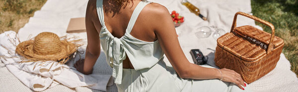 partial view of african american woman sitting near straw hat on blanket in park, summer picnic